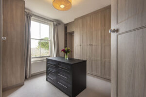 A walk-in wardrobe with a centre island dressing table with a window and wardrobe cupboards.